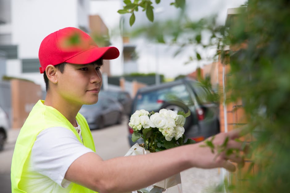 A delivery person wearing a red cap and a neon yellow vest is placing a bouquet of fresh white hydrangeas wrapped in white paper and tied with a ribbon onto a doorstep. The arrangement features vibran
