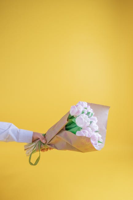 A hand holds a bouquet of fresh, pale pink tulips wrapped in light brown kraft paper with a green ribbon, set against a bright yellow background. The tulips have smooth, rounded petals and sturdy gree