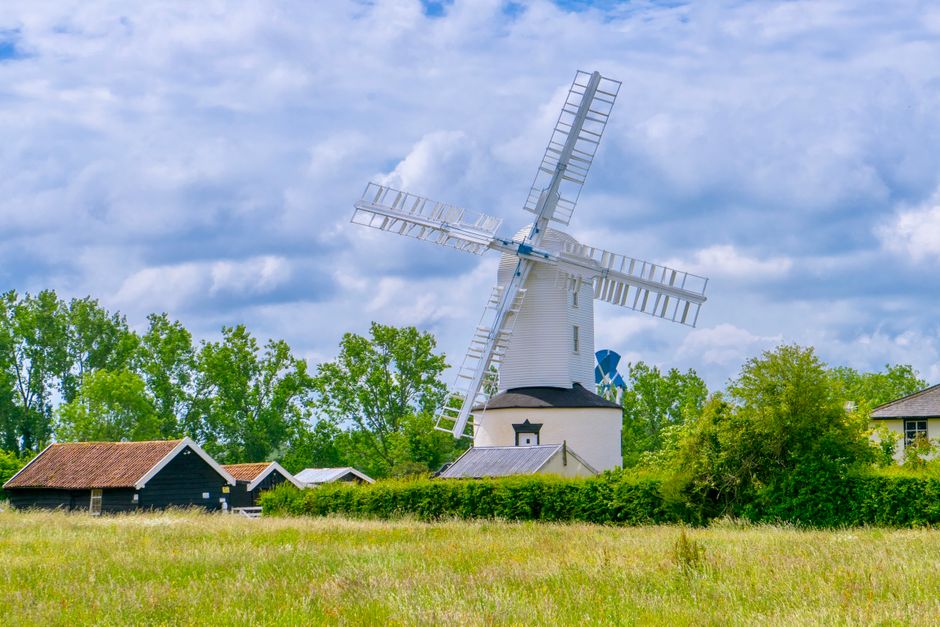 A white traditional windmill with four large sails stands prominently amidst lush green trees and a grassy field, with a few small farm buildings featuring red and gray roofs nearby. The sky above is