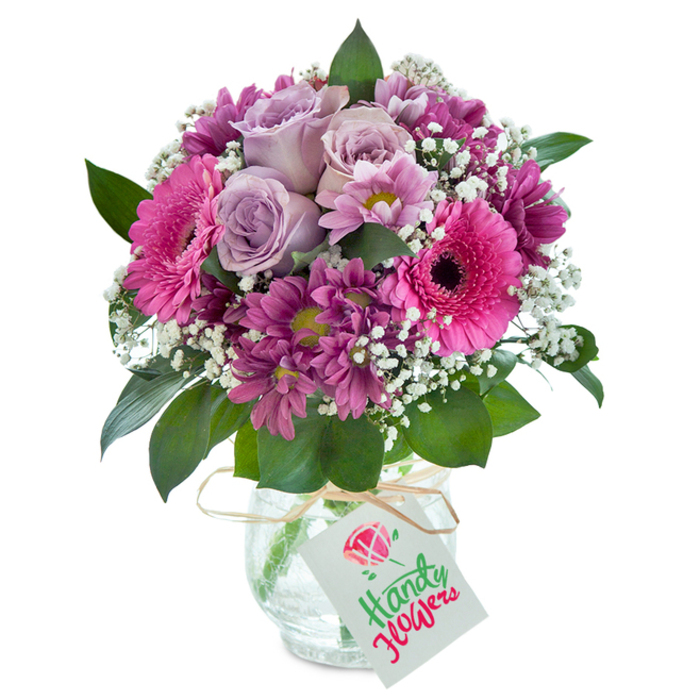 Eloquent and contemporary, this abundant floral bouquet finds its place against a clean, unadorned white backdrop, perfectly suiting Wennington's modern yet pastoral spirit. In the center, pale lavender roses delicately unfold, offering a gentle grace and wafting a suggestion of sweet fragrance. These are bordered by lively gerbera daisies in vivid fuchsia pinks-their wide, open faces exuding cheer reminiscent of local festivities along Wennington Road. The arrangement is texturally enriched by clusters of smaller, plush chrysanthemum blooms in lush magenta and plum, all highlighted by occasional yellow-green eyes. Sprays of pristine white baby's breath weave throughout the bouquet, imbuing it with airiness and clarity, as if reflecting sunlight through Wennington's leafy avenues. A selection of glossy, deep green leaves nestles among the blooms, grounding the composition and bringing out the vibrance of the palette. The flowers are gathered in a unique, rounded clear glass vase, its surface tinged with a delicate crackle that plays with the changing light. The vase's neck is tied with a rustic strand of raffia, holding a simple white card tag ready for a handwritten note or local greeting. This carefully considered bouquet from Wennington Florist makes a striking gift for any milestone-a birthday, heartfelt thank-you, or a moment to simply brighten a neighbour's day-delivered fresh within Wennington and surrounding villages.
